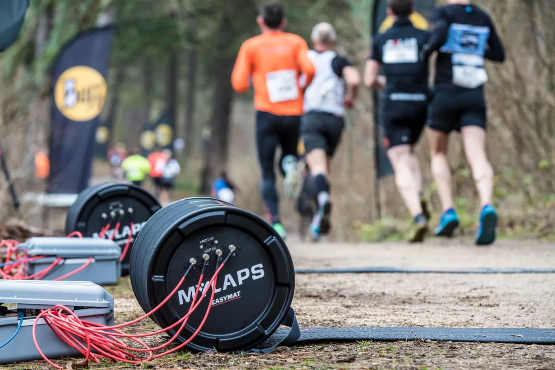 Black and yellow MYLAPS timing mats laid across a forest trail at a race, with runners moving away into the distance, used to read RFID transponders as competitors pass over them.